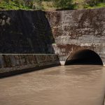 Galeria de drenagem de concreto com água escura escoando para um túnel, entre paredes cobertas de musgo.