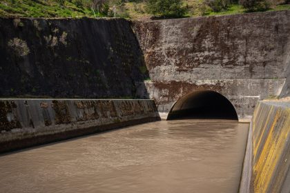 Galeria de drenagem de concreto com água escura escoando para um túnel, entre paredes cobertas de musgo.