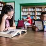 Crianças leem livros sentadas a uma mesa em uma biblioteca escolar, cercadas por estantes.
