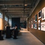 Interior de uma galeria de arte com obras emolduradas e visitantes sentados sob iluminação aconchegante.