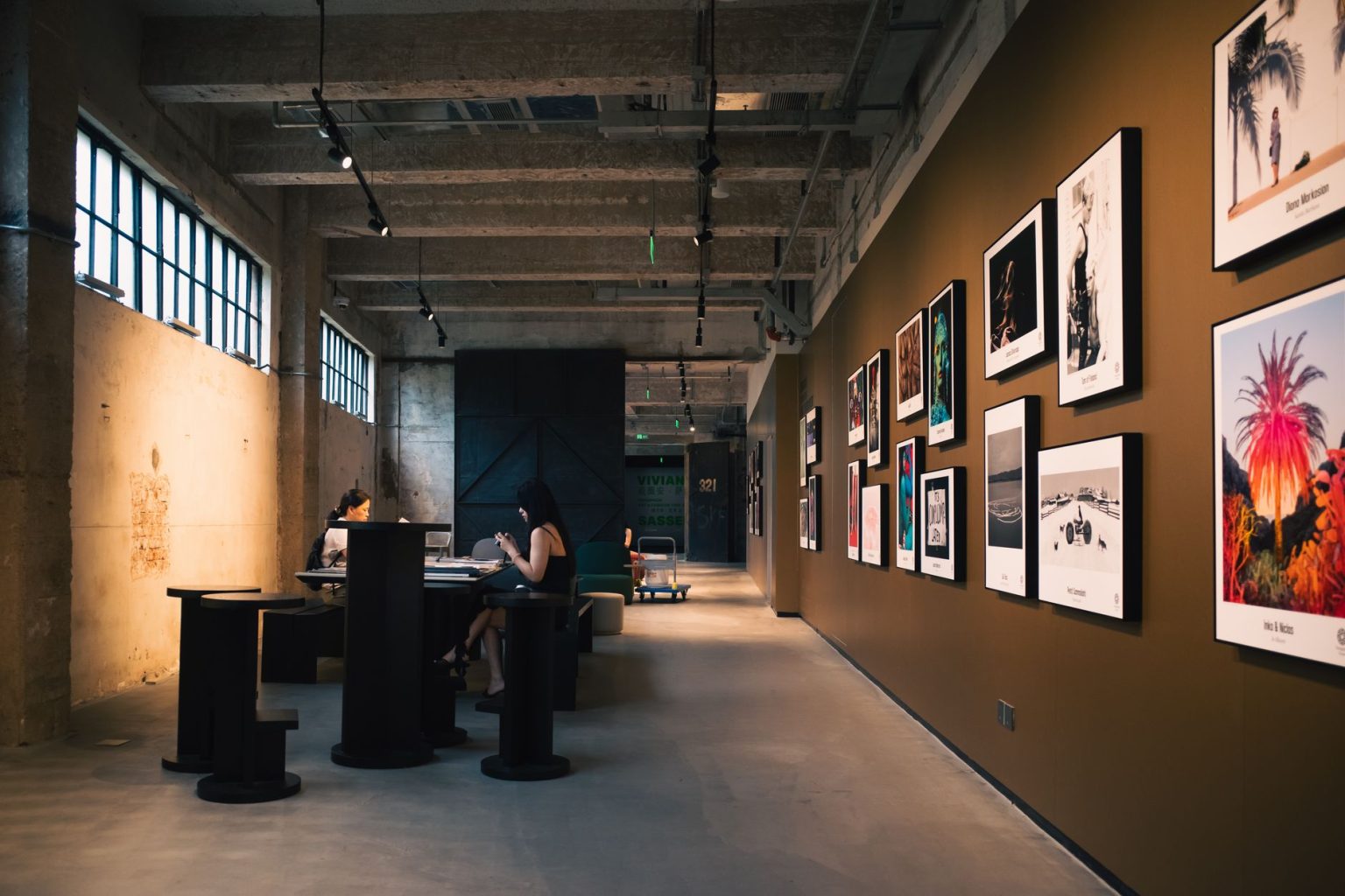 Interior de uma galeria de arte com obras emolduradas e visitantes sentados sob iluminação aconchegante.