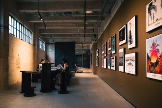 Interior de uma galeria de arte com obras emolduradas e visitantes sentados sob iluminação aconchegante.