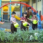 Crianças aprendem a regar plantas com a orientação de uma professora em um espaço verde da escola.