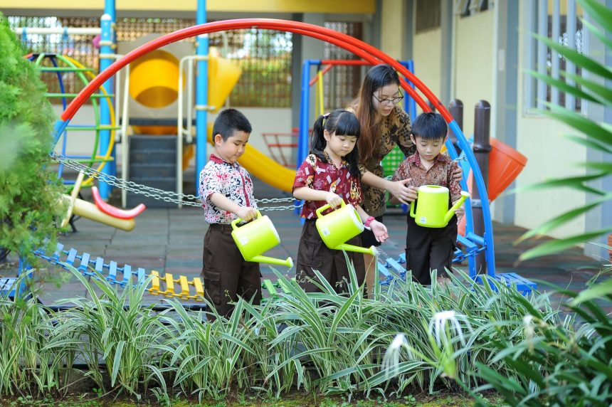 Crianças aprendem a regar plantas com a orientação de uma professora em um espaço verde da escola.